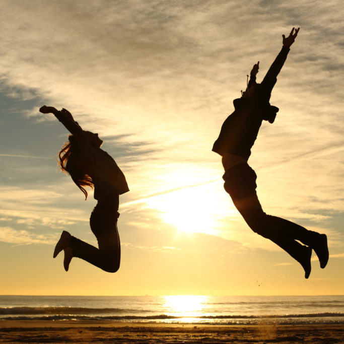 Couple or friends silhouette jumping on the beach at sunset with a warmth light and the sun in the middle